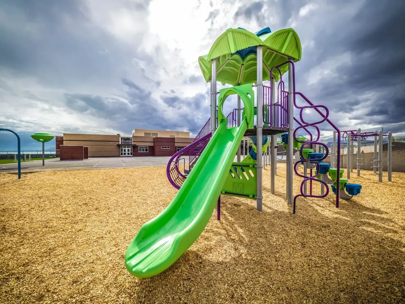 Playground with a green slide and climbing structure on wood chips, with the school building in the distance