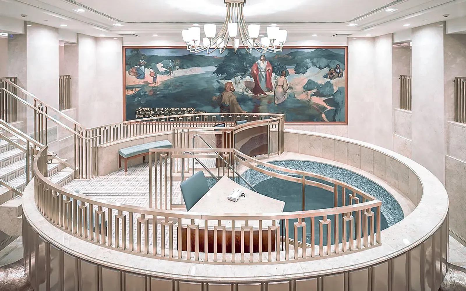 Baptistry with a circular font ringed by a railing, a large mural on the wall behind and a chandelier overhead
