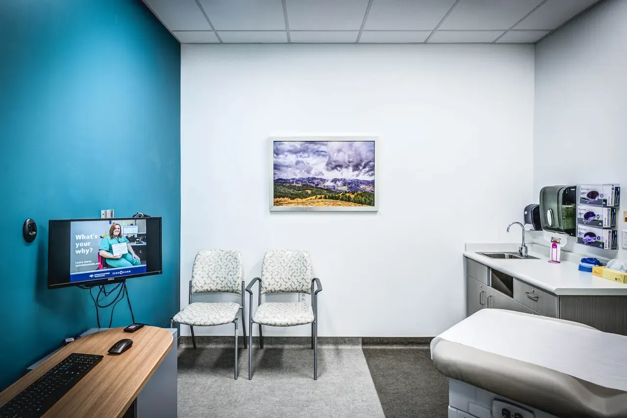 Exam room with a teal wall, two chairs, an exam table, a sink and a framed landscape.