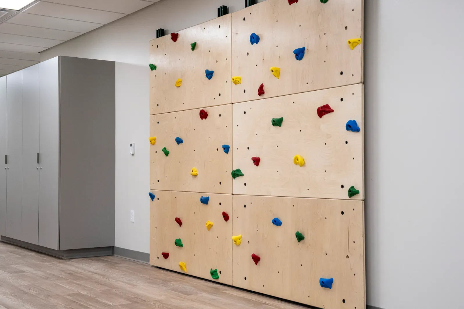 Wall-mounted climbing panels with colored holds beside gray storage cabinets.