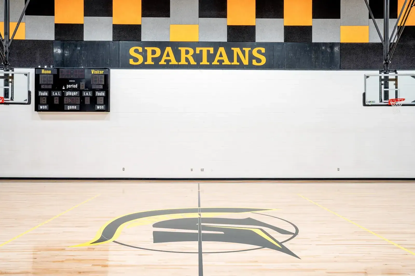 Gym wall with SPARTANS lettering, a scoreboard and a Spartan helmet logo at center court.