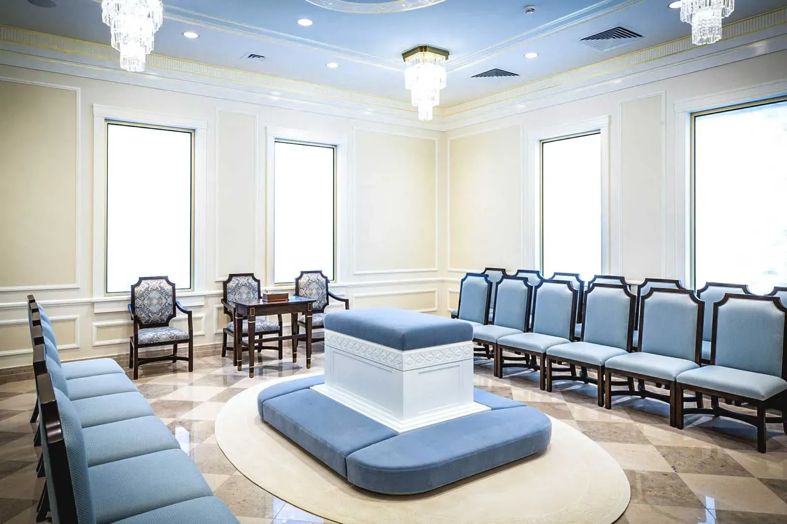 Room with a central blue-cushioned altar, blue upholstered chairs and tall windows.