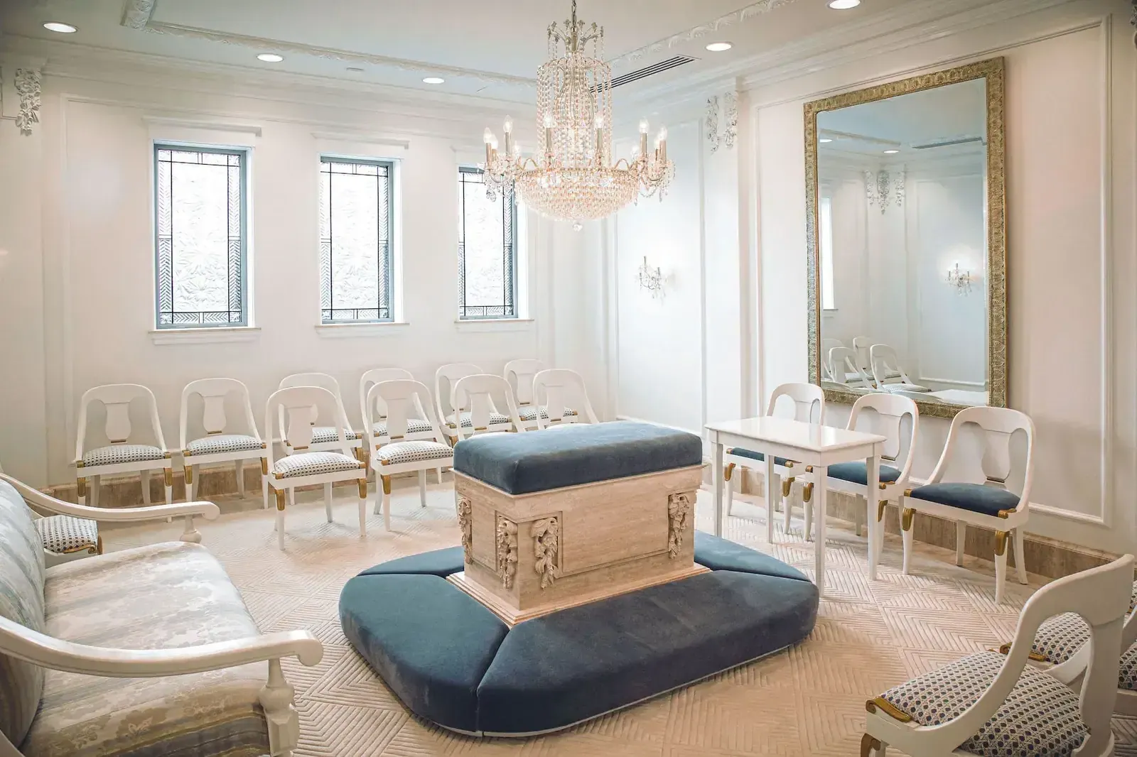 Room with a blue upholstered altar at the center, white chairs around it, a chandelier and a gold-framed mirror.