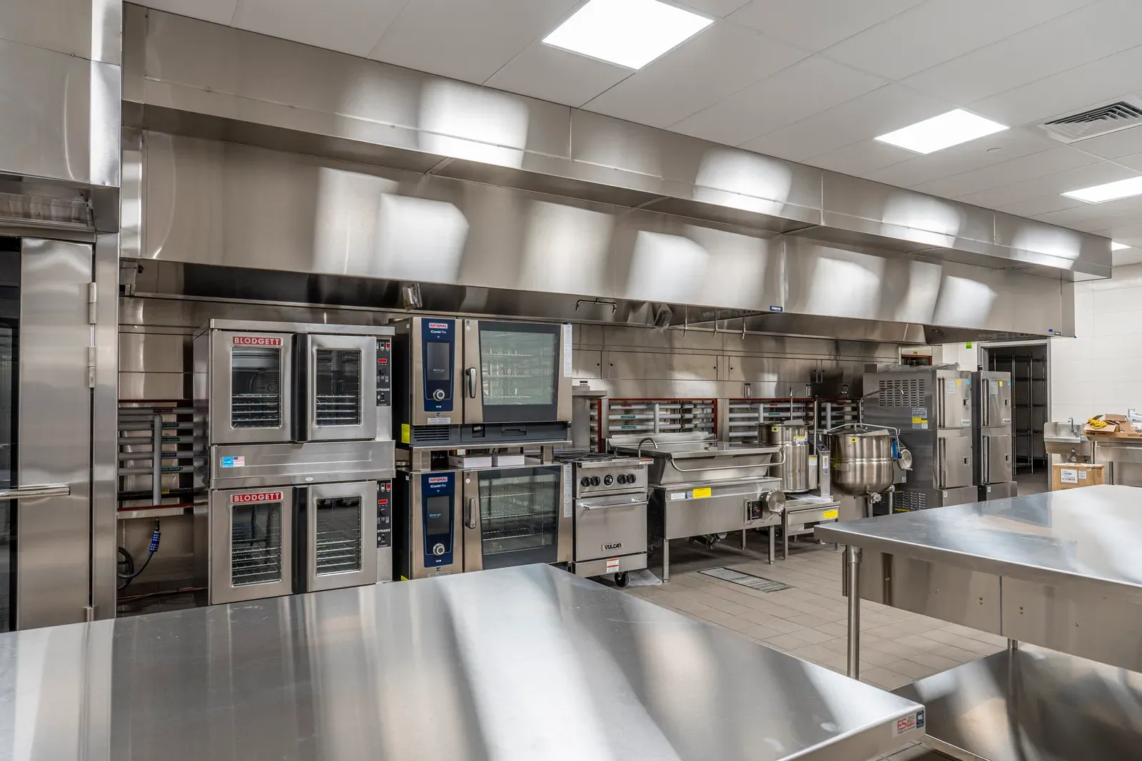 Kitchen with convection ovens, a steam kettle, a range under a hood and stainless steel prep tables.