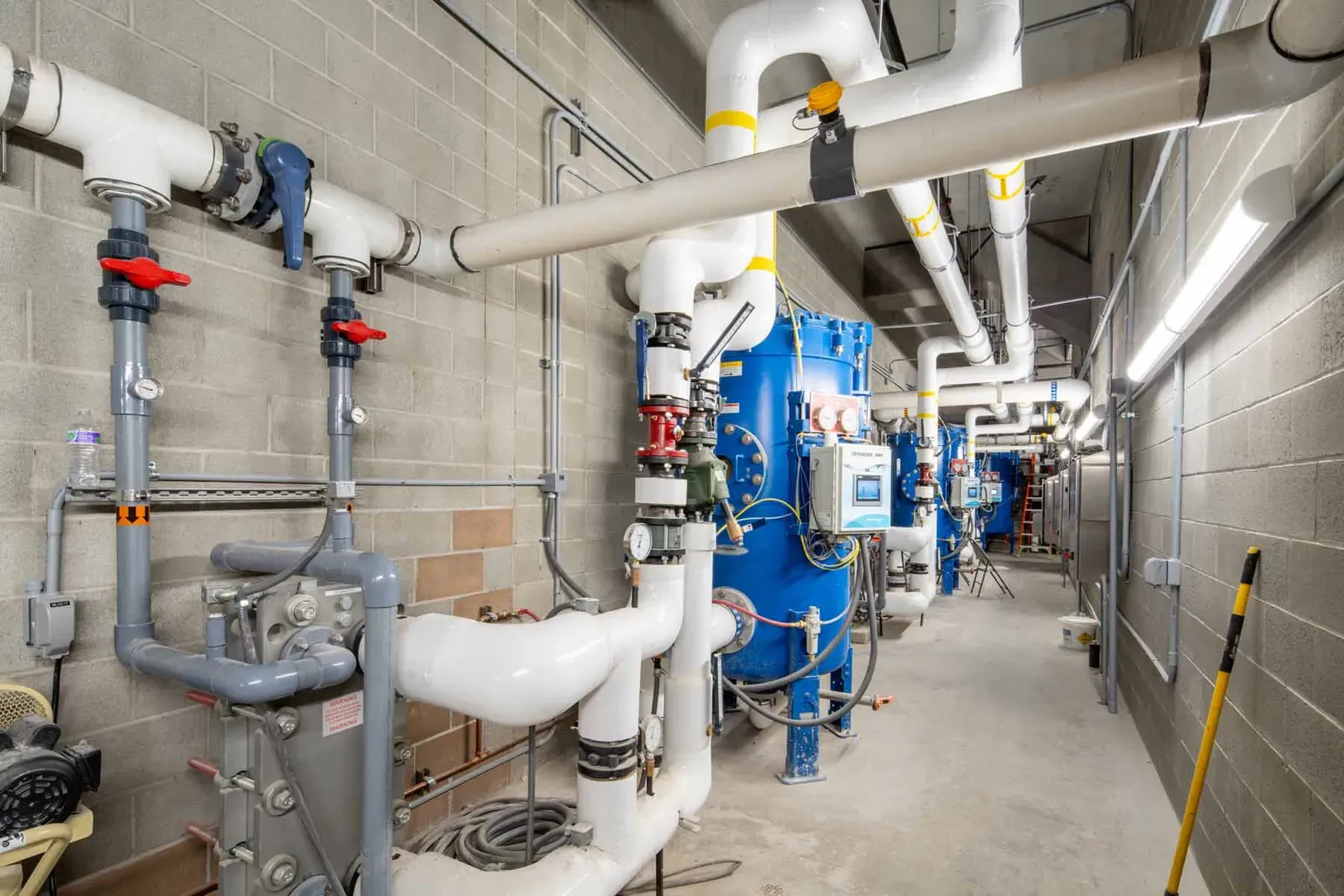 Pool mechanical room with white and gray piping and blue filter tanks along a block wall.