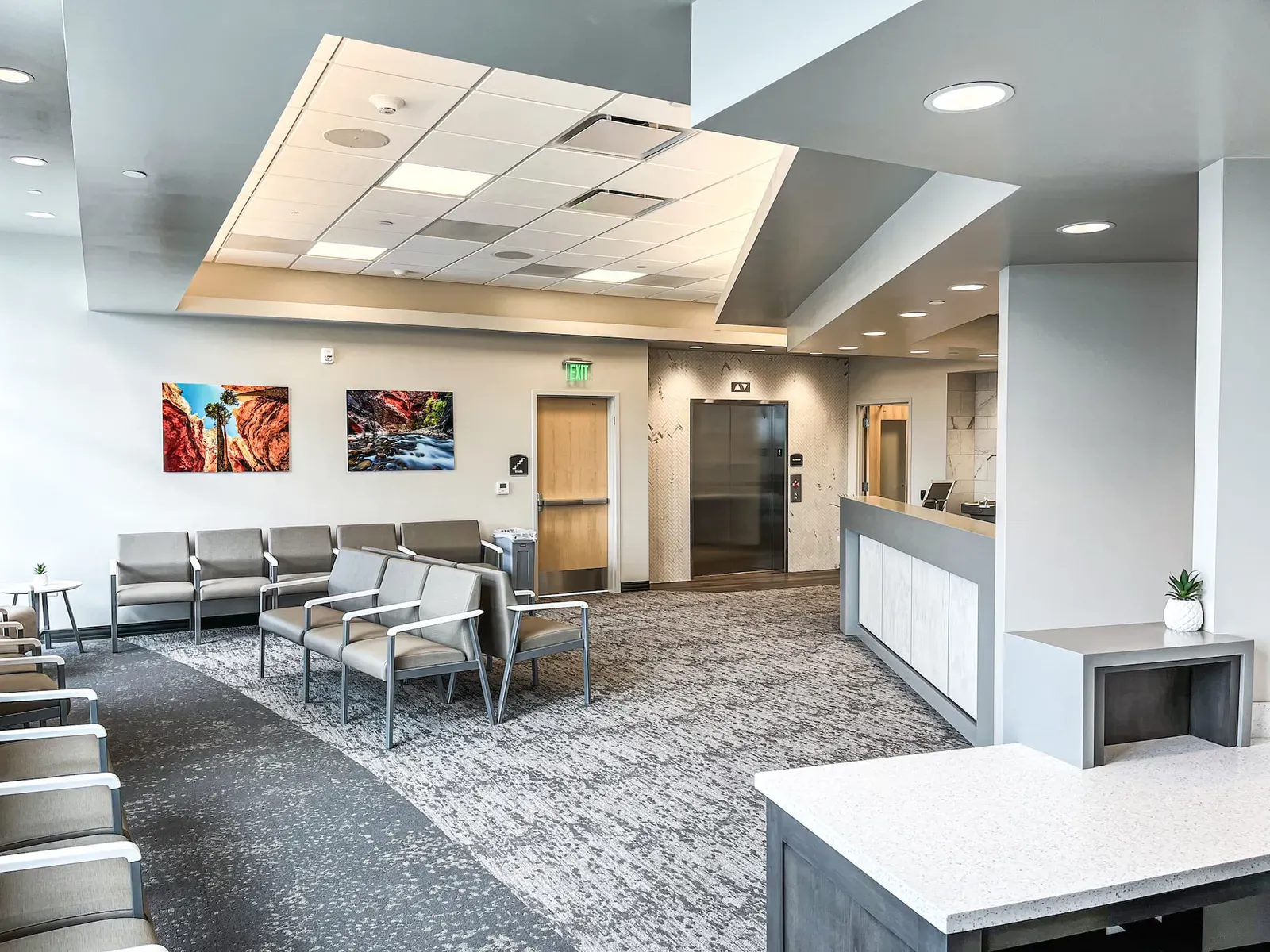 Waiting area with gray chairs and red rock landscape art beside a reception counter and elevator