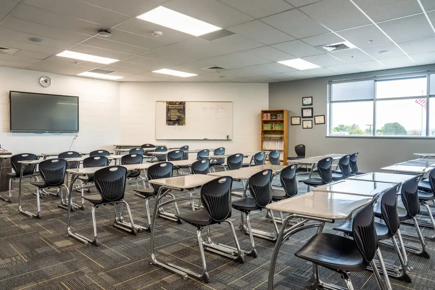 Classroom with rows of student desks, black chairs, a wall-mounted display and a window along one wall.