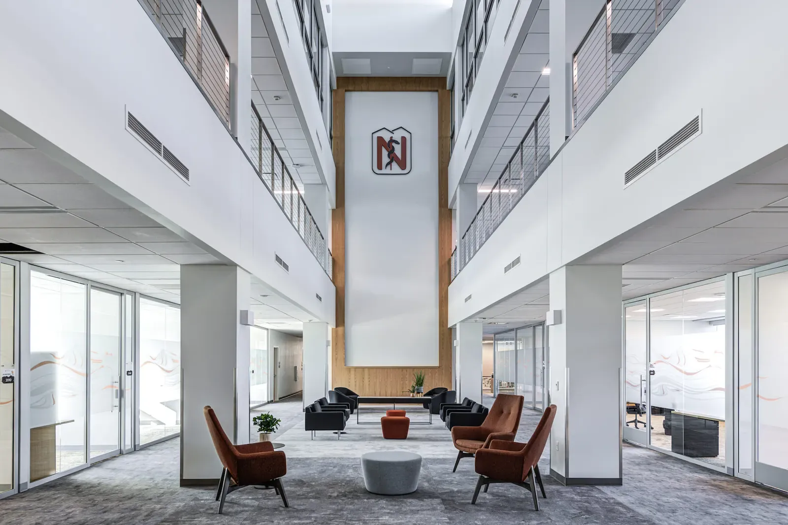 Atrium with the college logo on a tall wood-framed wall, lounge seating and glass-walled rooms on both sides.