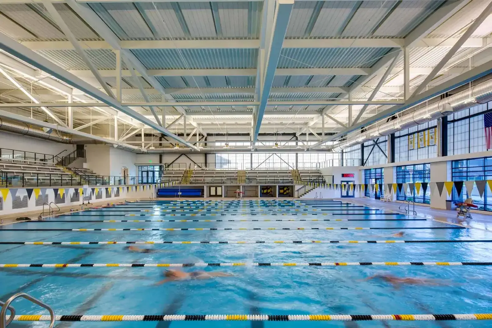 Competition pool with lane lines, spectator seating along the far wall and a steel truss roof overhead.