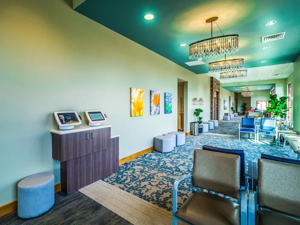 Waiting area with check-in kiosks on a wood cabinet, ottomans, a teal ceiling and crystal chandeliers.