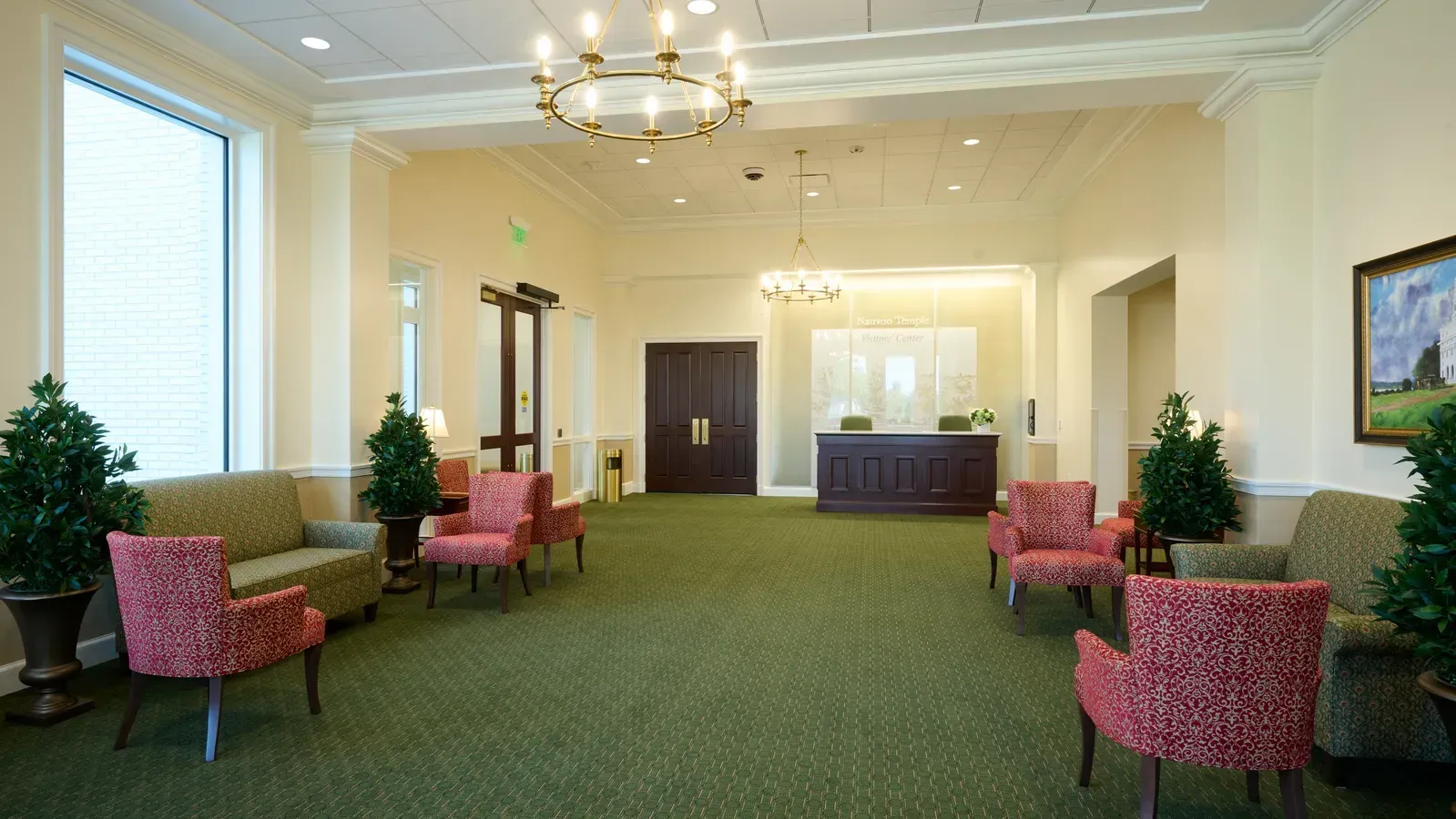 Lobby with a reception desk, red patterned armchairs, green sofas, two chandeliers and a landscape painting on the wall.