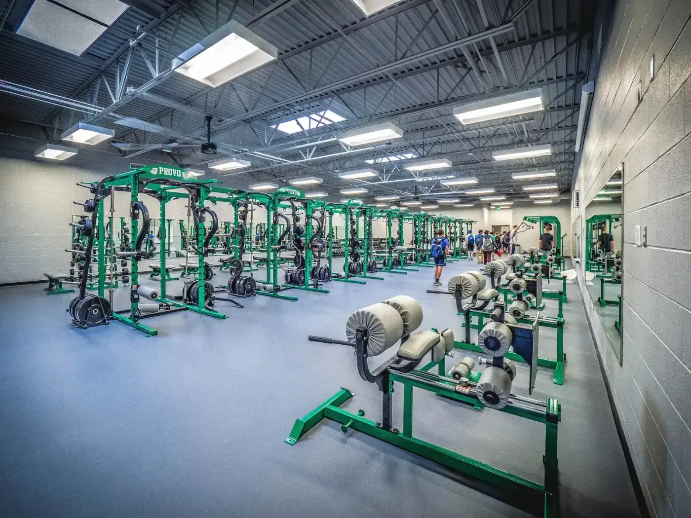 Weight room with rows of green power racks and weight machines on a blue rubber floor