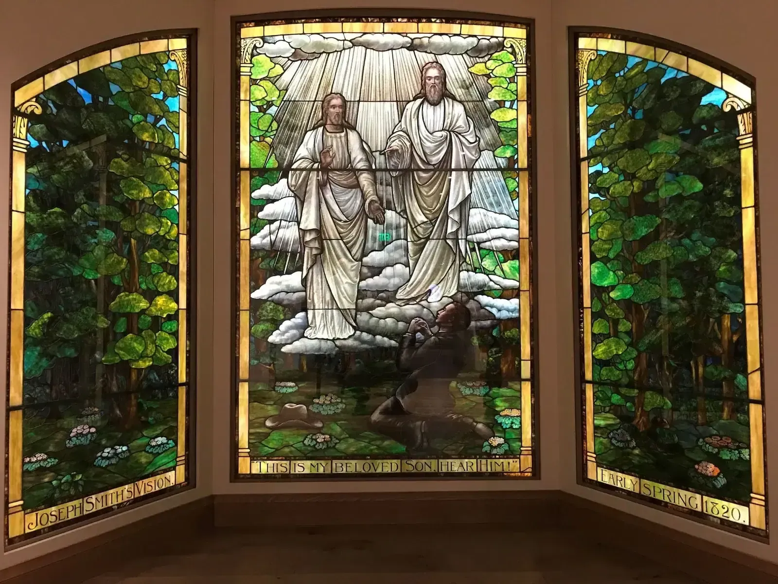 Three-panel stained glass window showing a forest and two robed figures among clouds