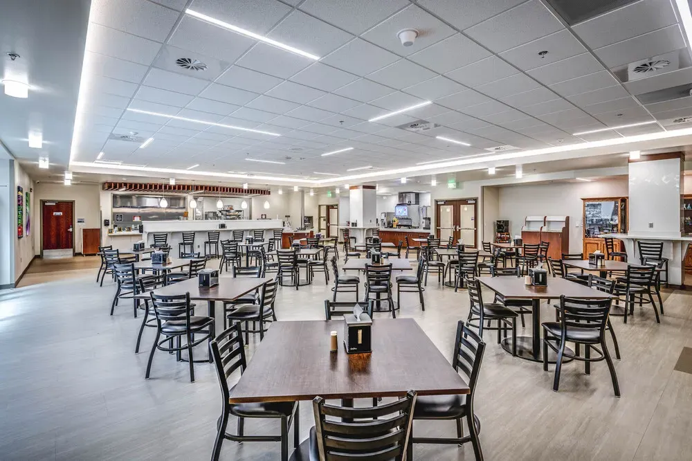 Cafeteria dining room with dark wood tables and black chairs, and the serving counter at the back