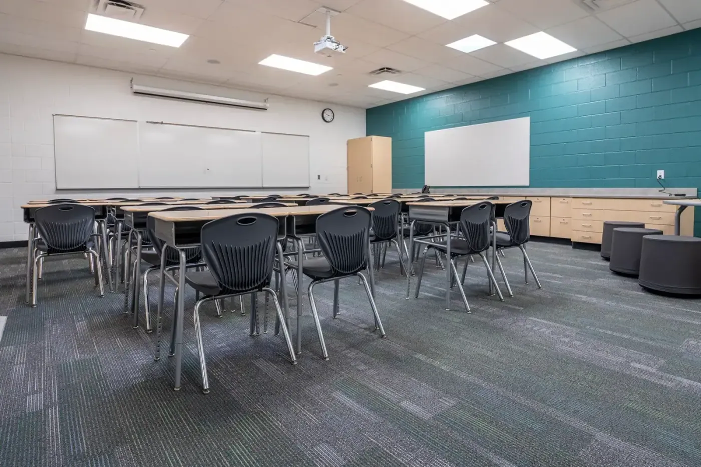 Classroom with student desks, black chairs, whiteboards, a teal accent wall and wood cabinets.