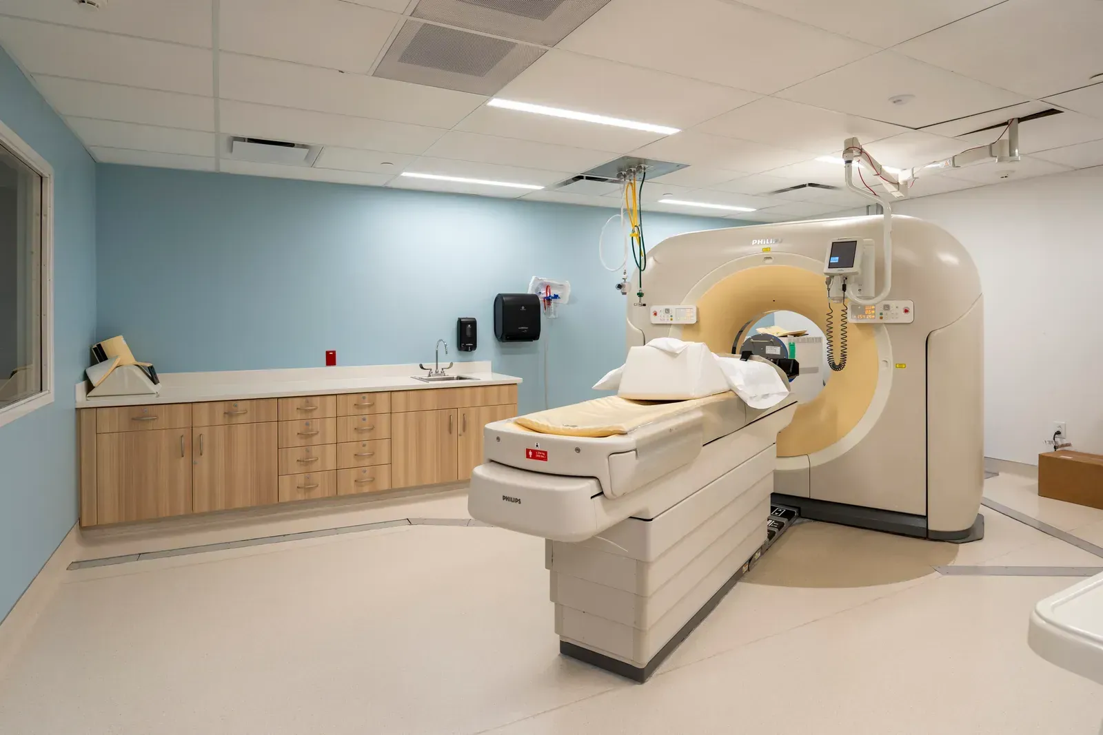 CT scanner room with a patient table, light blue walls and wood cabinets.
