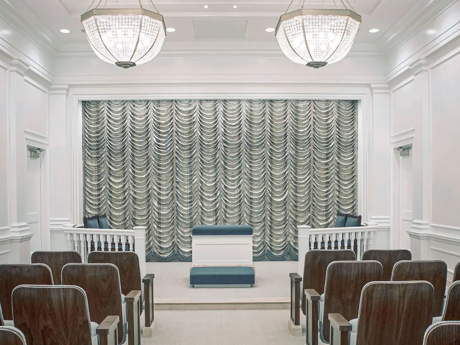 Room with a draped gray curtain behind a raised stand, wood-backed seating and two crystal chandeliers.