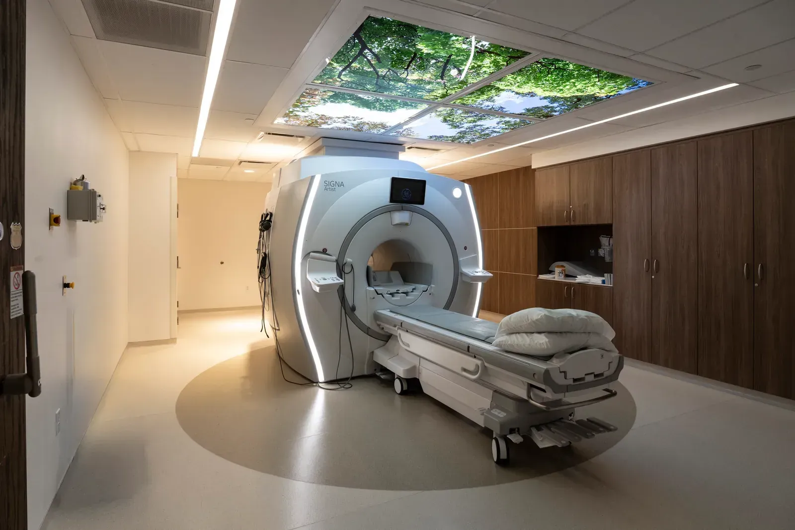 MRI scanner room with a tree-image ceiling panel and wood cabinets along one wall.