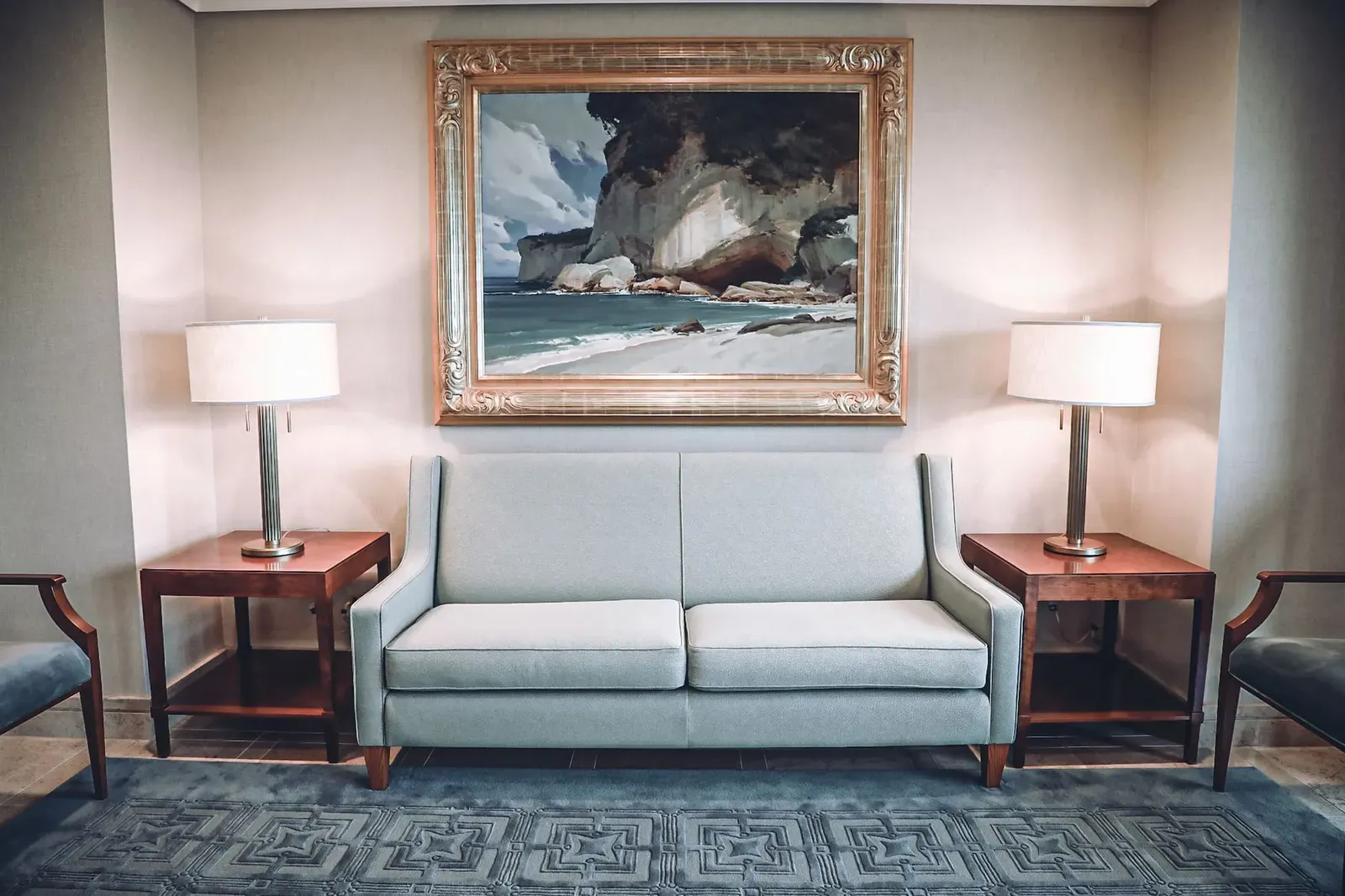 Seating area with a pale blue sofa between two lamps under a large framed painting of a rocky seashore