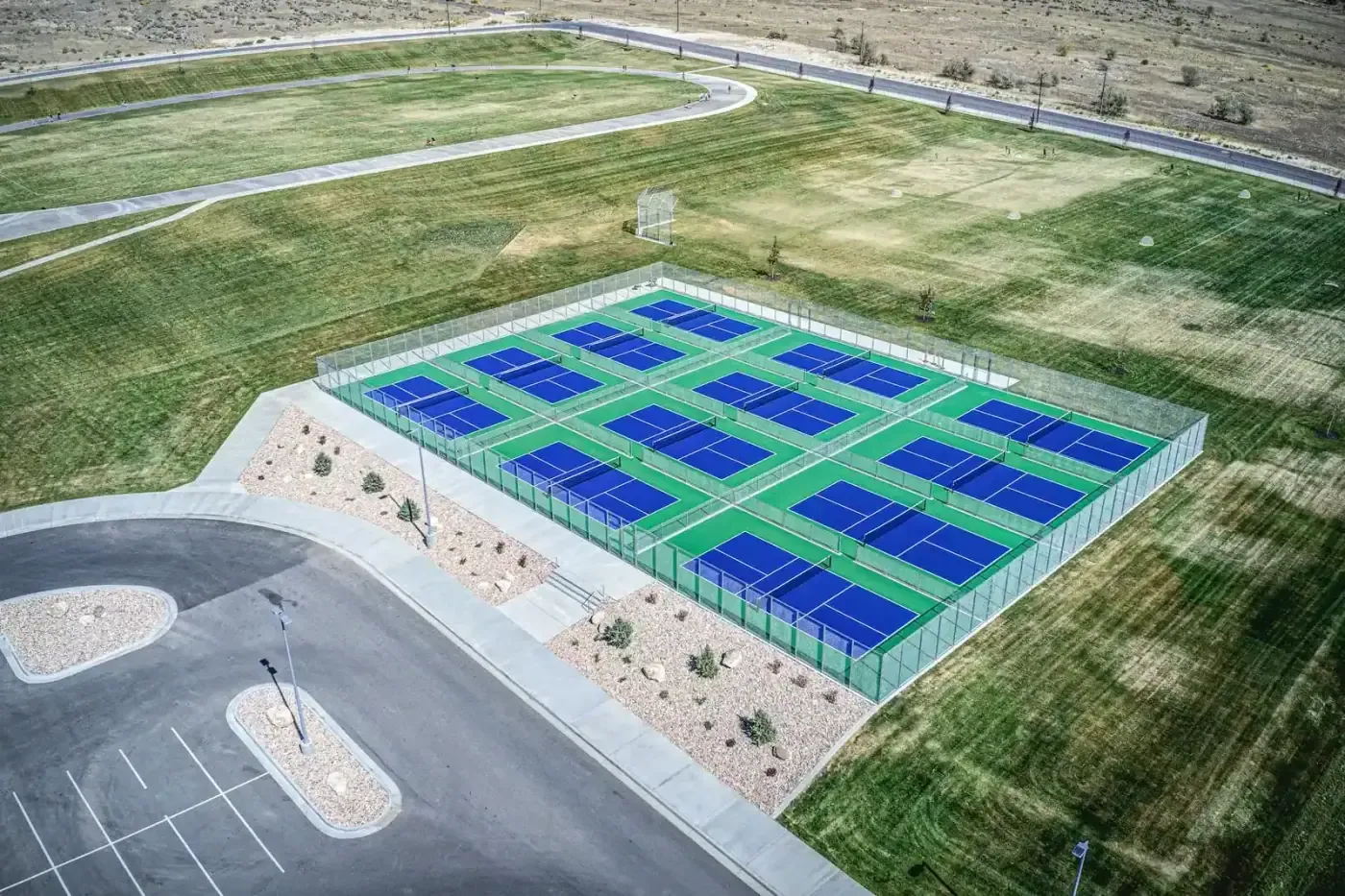 Aerial view of fenced blue and green sports courts next to a parking lot and open fields.