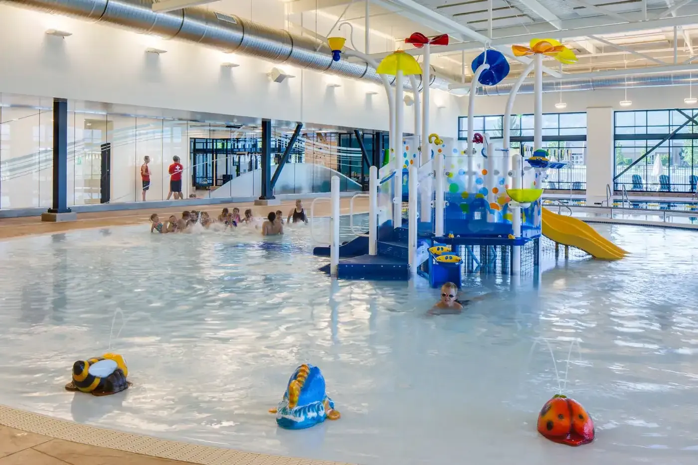 Zero-entry recreation pool with children playing near a water play structure and slide.