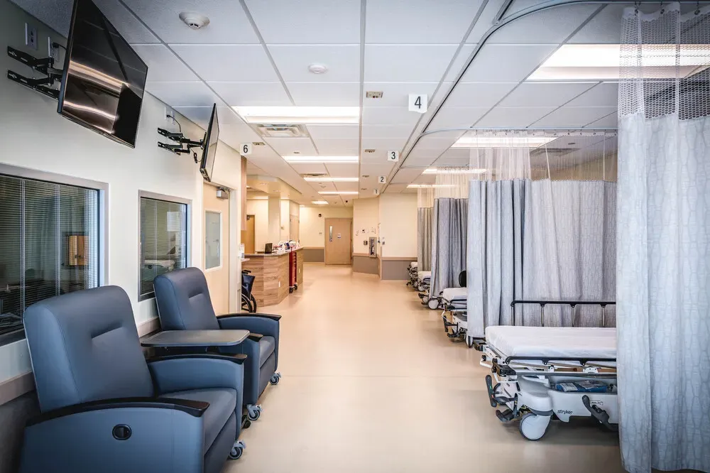 Recovery bay corridor with two recliner chairs, wall-mounted TVs, and curtained stretcher bays.