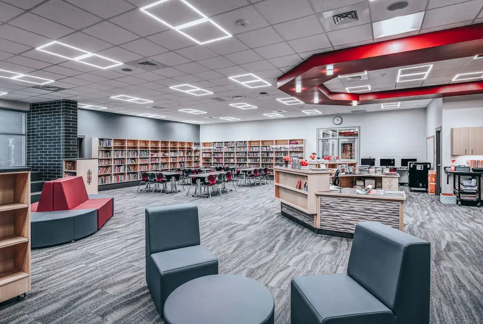 Library seating area with gray lounge chairs, a red bench and long rows of bookshelves