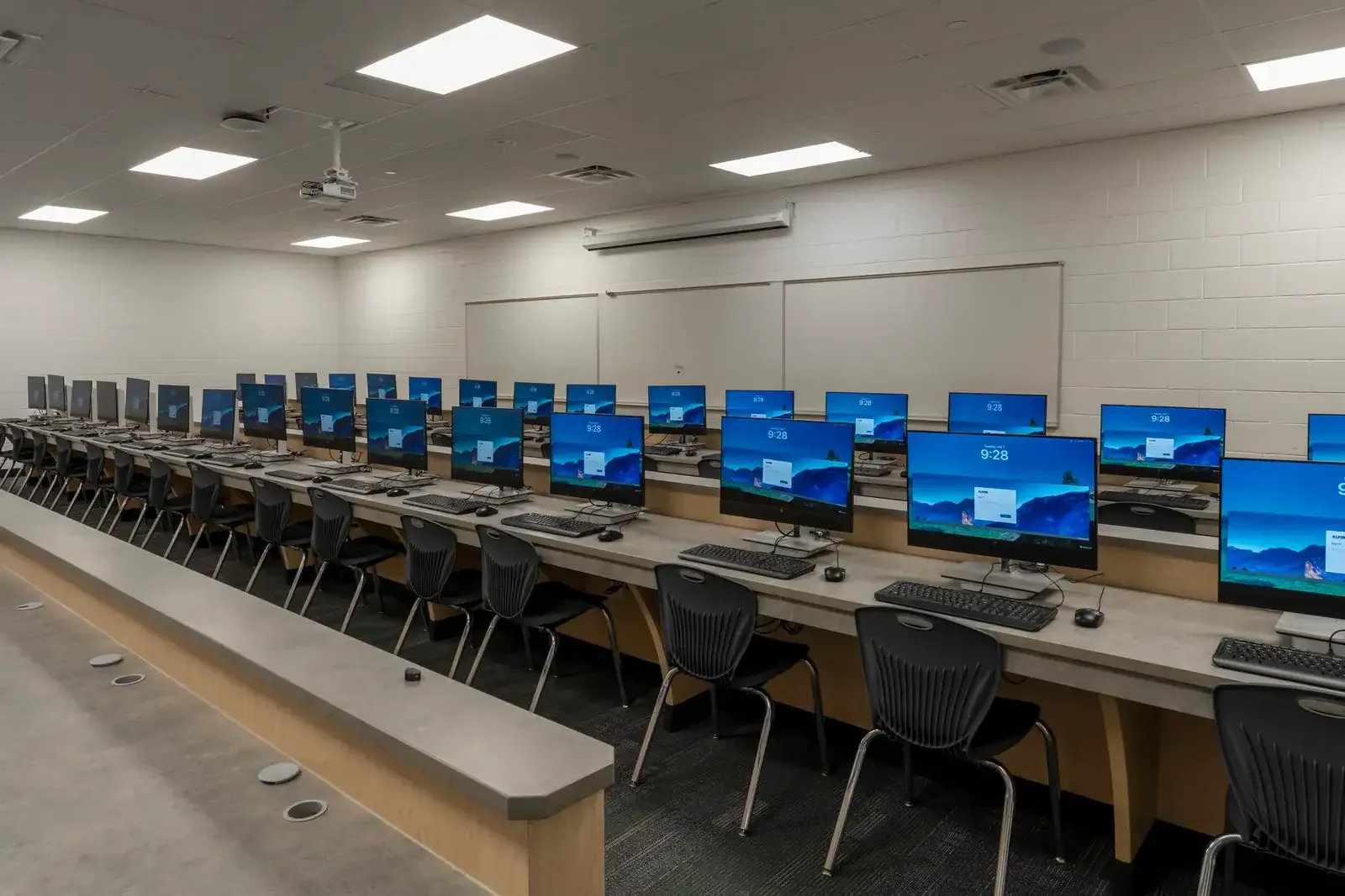 Computer lab with a long counter of monitors, black chairs and whiteboards on a white block wall.