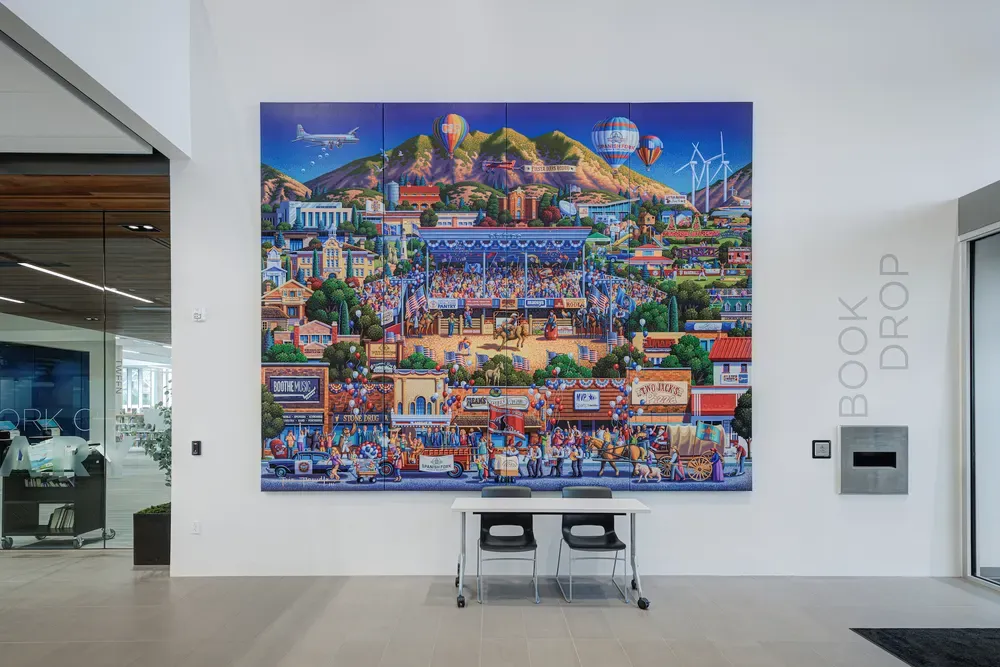 Large illustrated mural of a town festival on a lobby wall above a table, next to a Book Drop sign