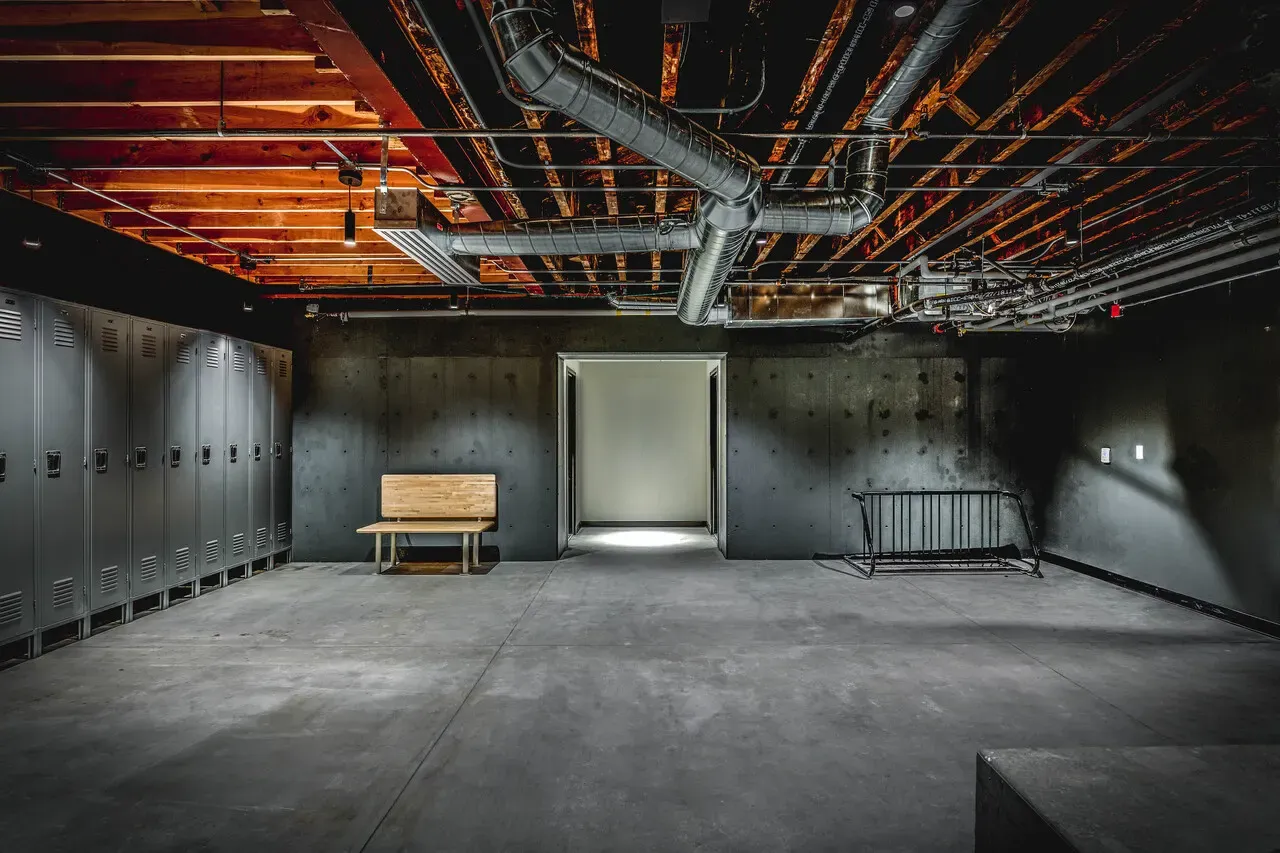 Room with a row of gray lockers, a wooden bench and exposed ductwork overhead.