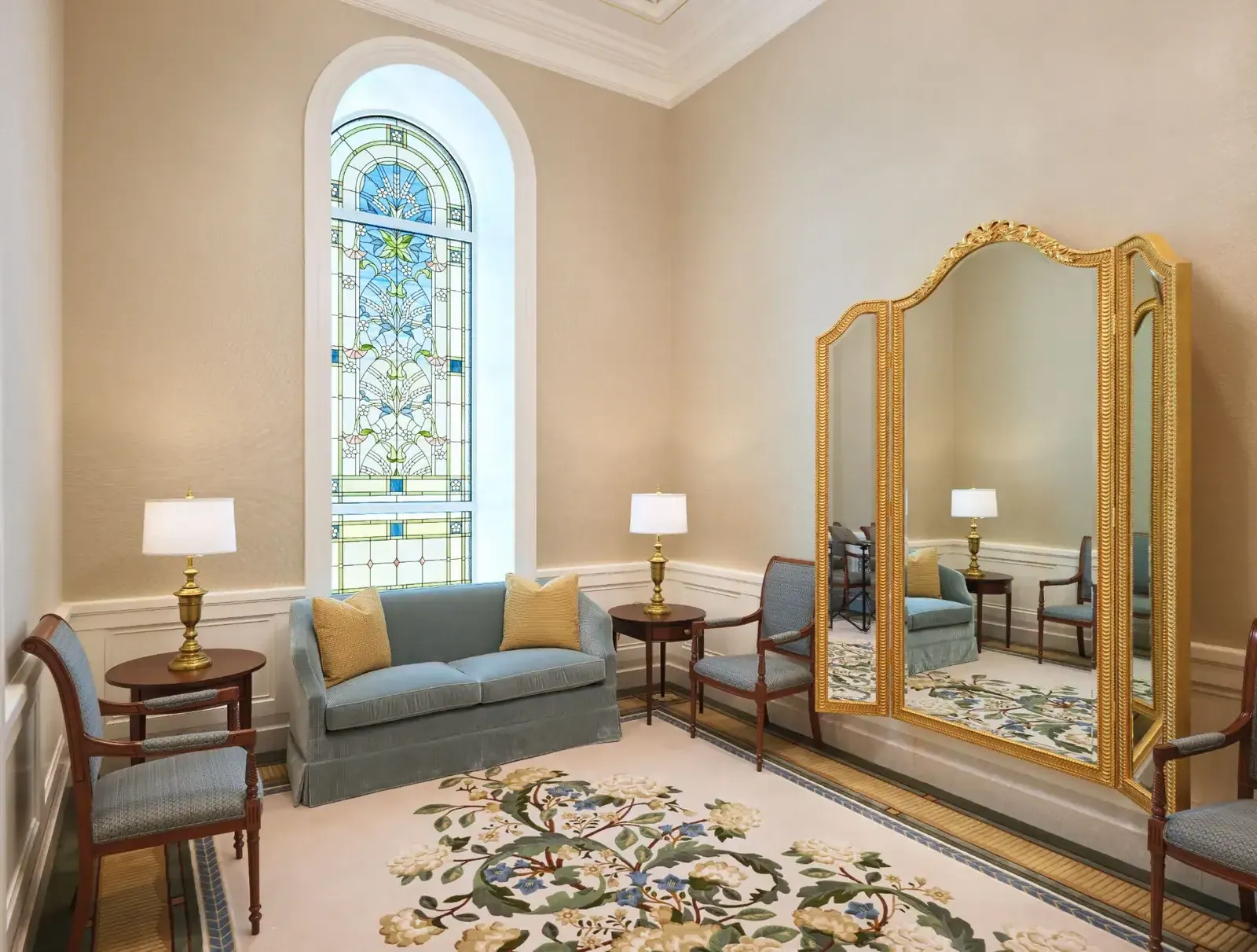 Room with a tall stained-glass window, a blue sofa, table lamps and a gold-framed three-panel mirror over a floral rug