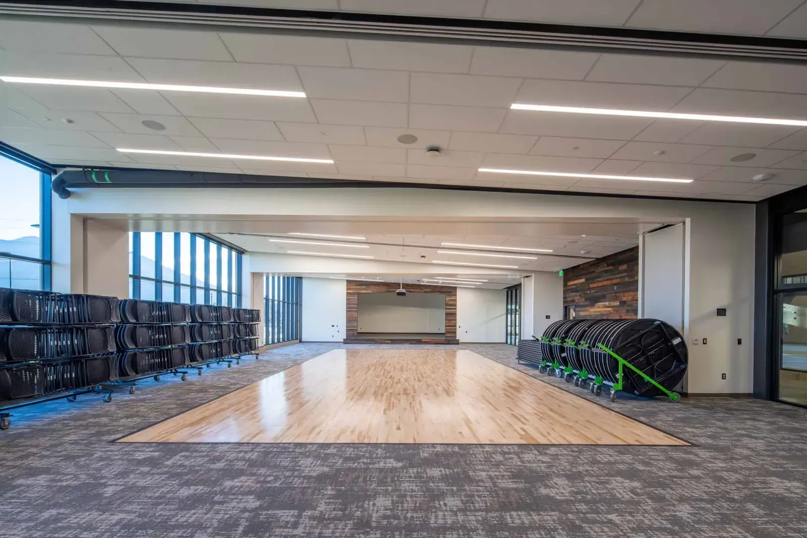 Multipurpose room with a wood floor at the center, carpet around it, stacked round tables on carts and tall windows.