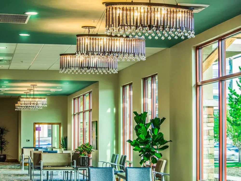 Crystal chandeliers hanging from a teal ceiling above waiting area seating, with tall windows and a potted plant at right.