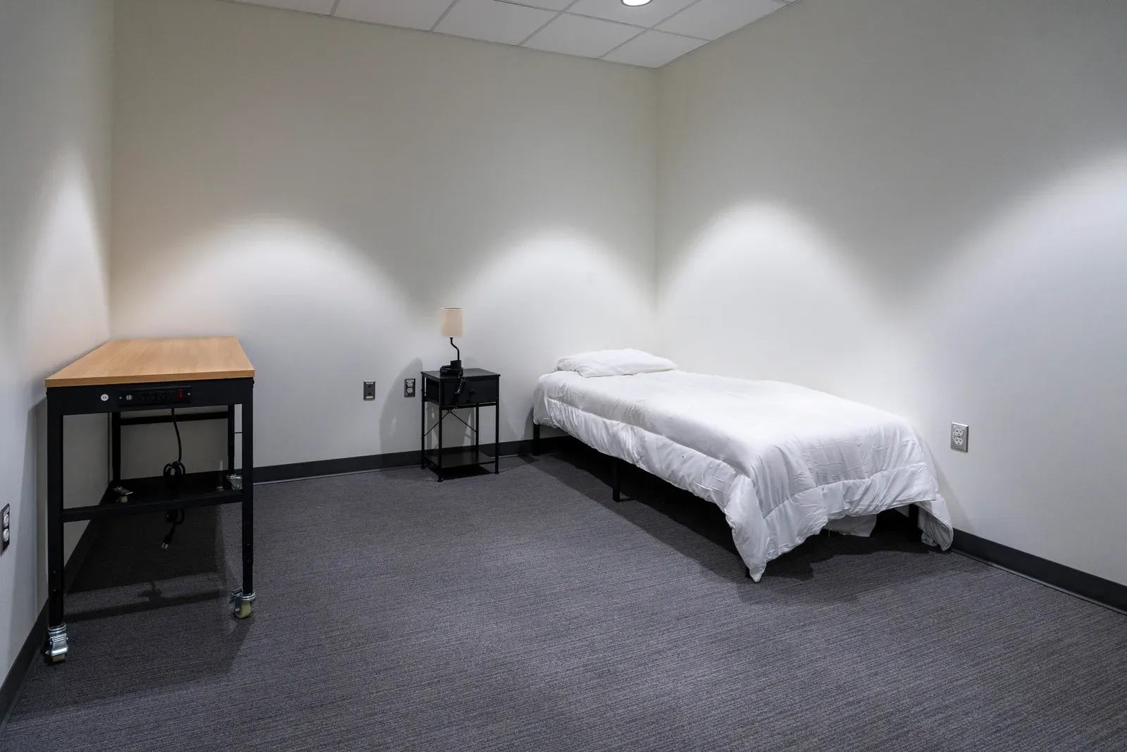 Crew quarters room with a single bed, a nightstand with a lamp and a rolling work table.