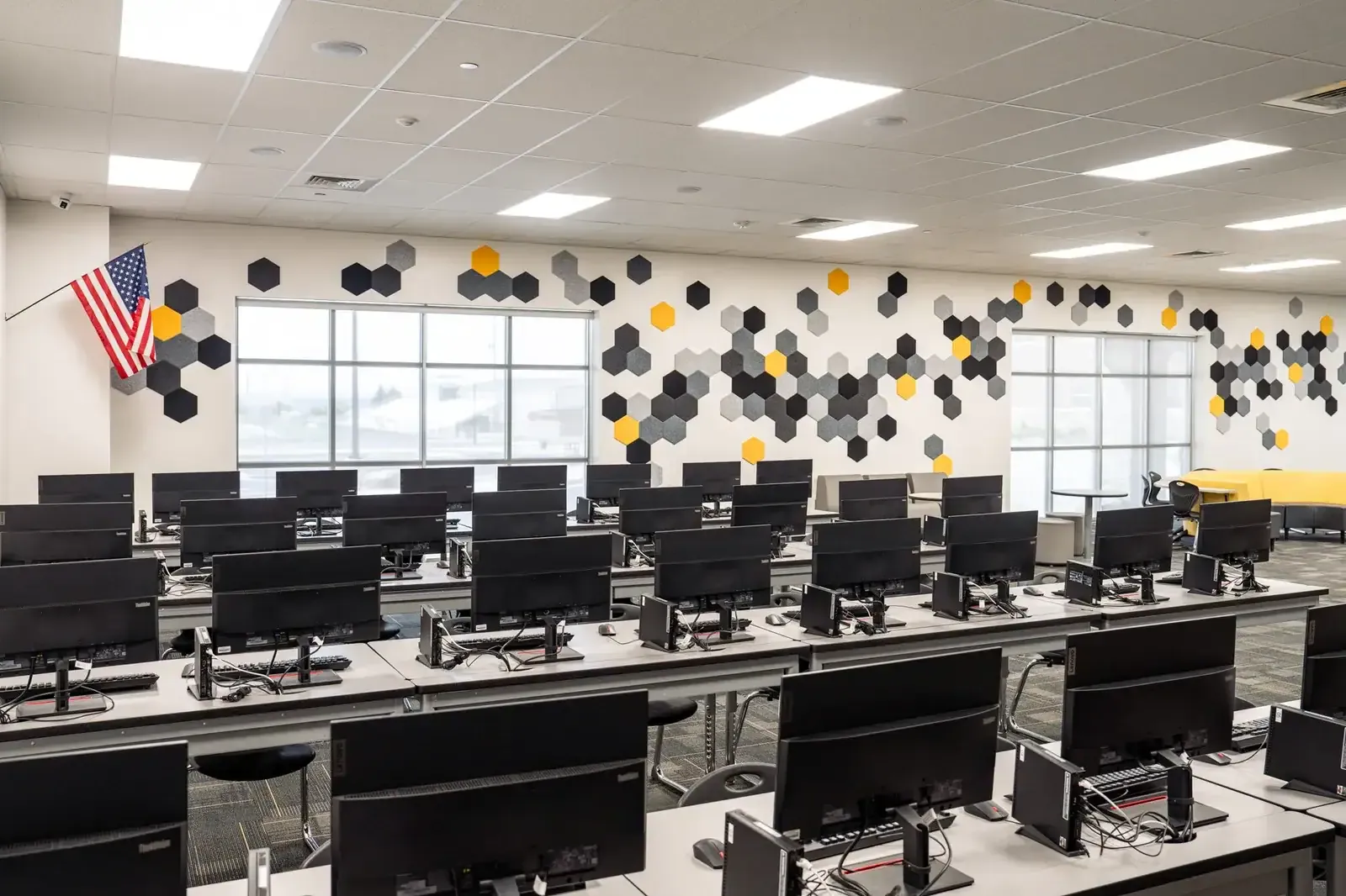 Computer lab with rows of monitors, a wall of hexagon acoustic panels in black, gray and gold, and two window walls.
