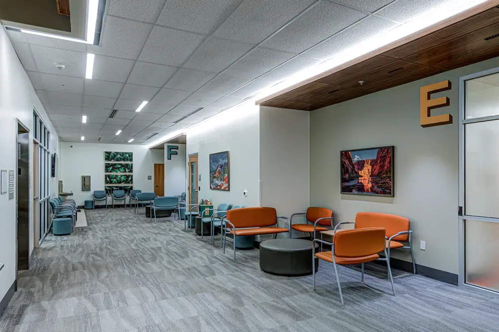 Corridor with orange and teal waiting chairs, a wood ceiling band and lettered wayfinding signs.