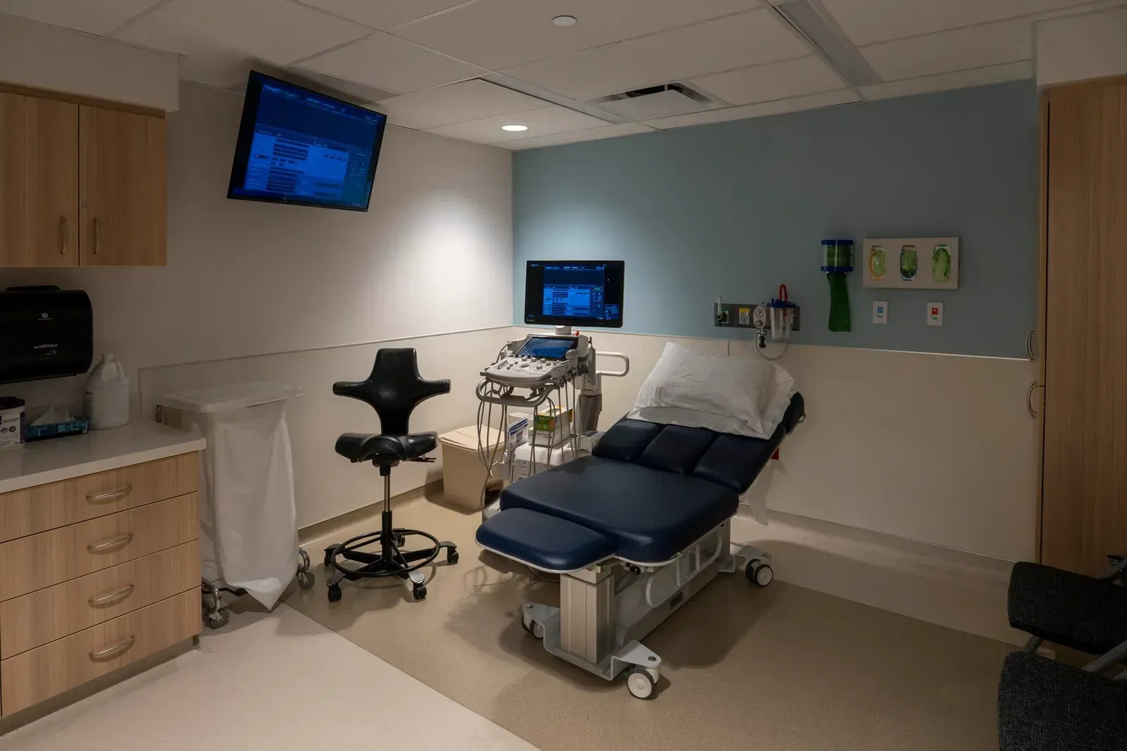 Ultrasound exam room with an exam table, an ultrasound cart, a wall-mounted screen and cabinets.