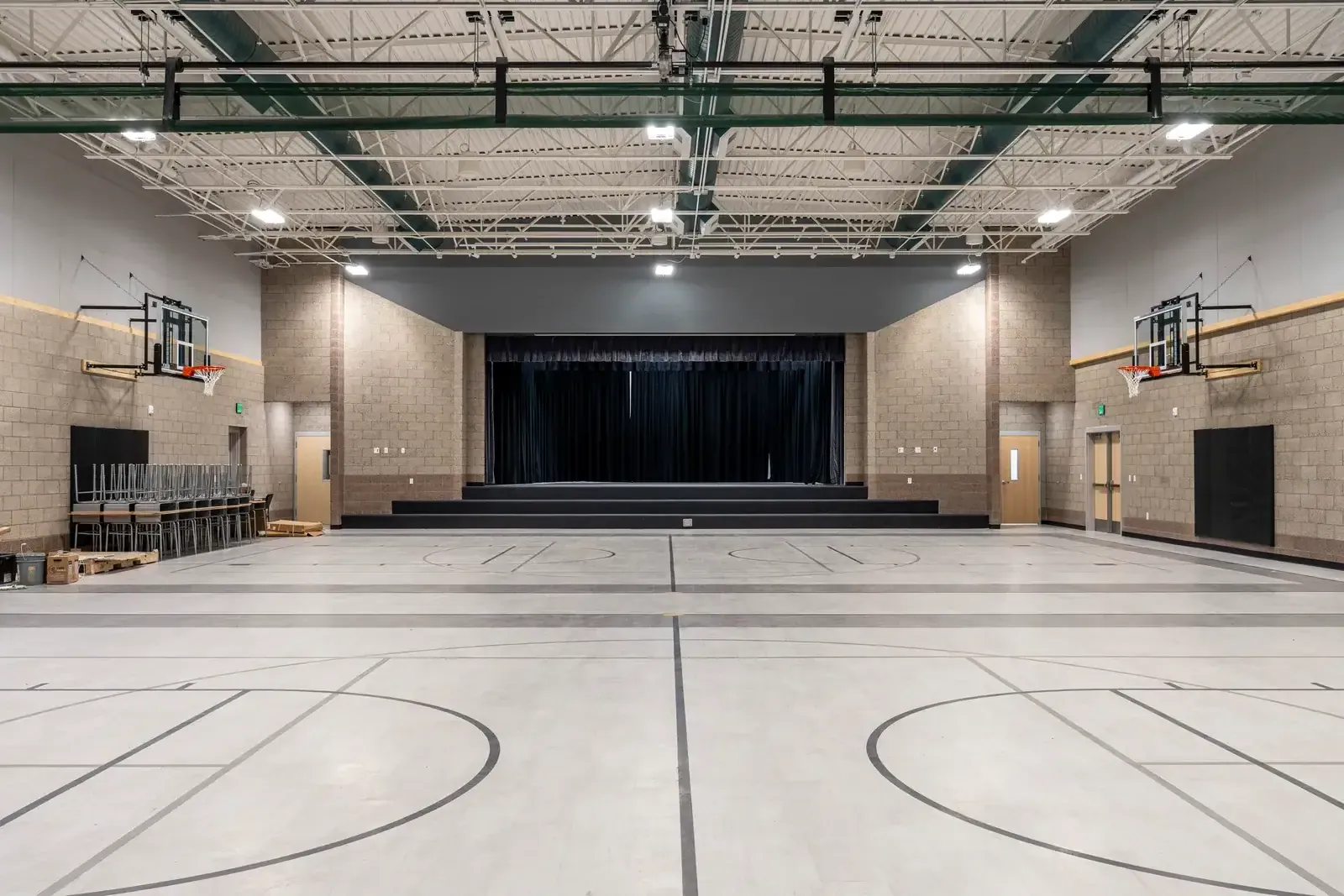 Gym with a gray floor and court lines, wall-mounted hoops and a stage with a dark curtain at the far end.