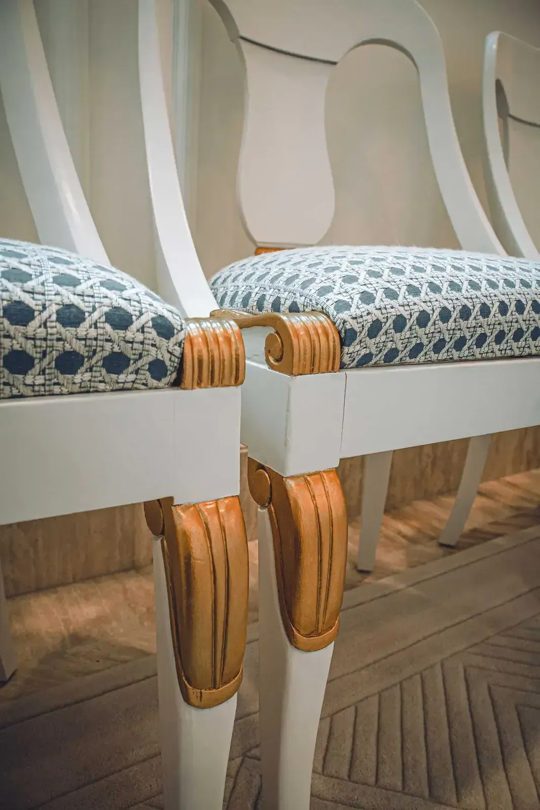 Close view of white chairs with woven blue seat fabric and gilded carved details on the legs.