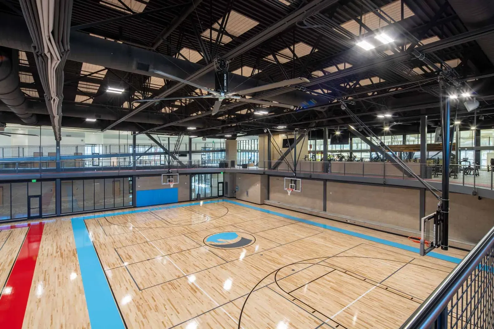 Gymnasium with a wood basketball court marked with blue lines and an upper-level walkway overlooking it.