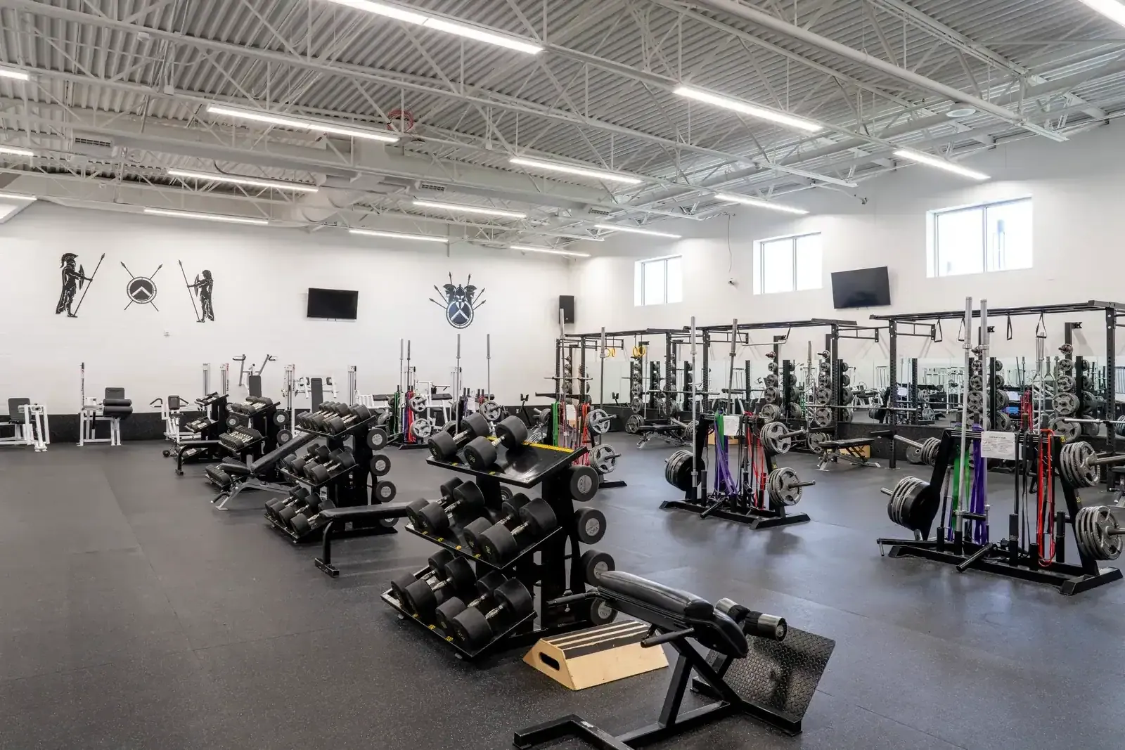 Weight room with racks, dumbbell stands and benches on a black rubber floor under a steel roof structure.