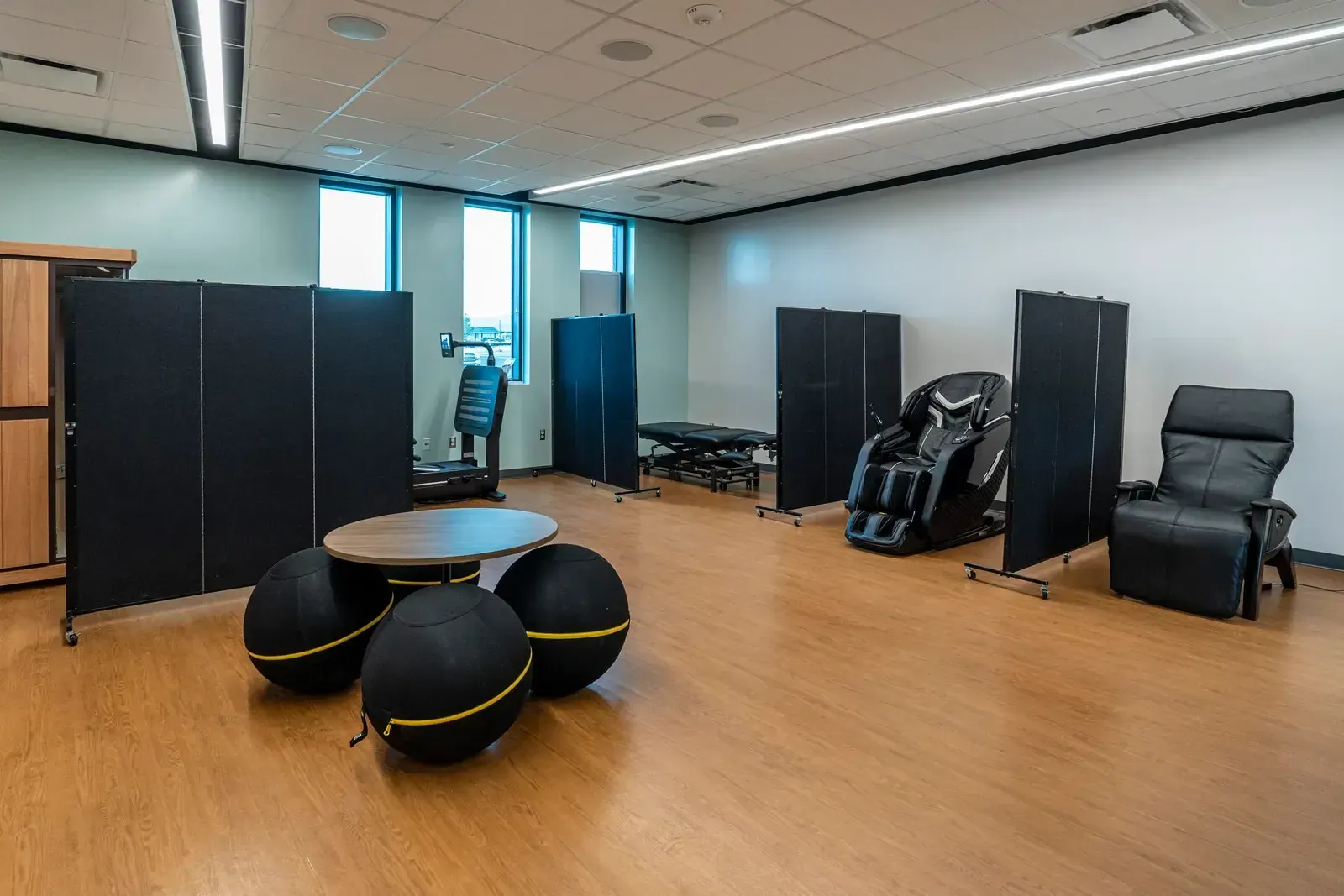 Wellness suite with massage chairs, treatment tables, privacy screens and exercise-ball seating around a small table.