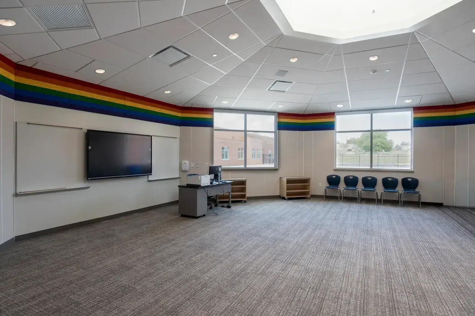 Classroom with a wall-mounted display, whiteboards, windows and a rainbow-colored band along the top of the walls.