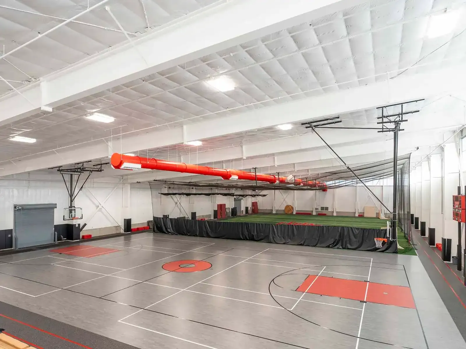 Indoor practice gym with a basketball court in the foreground and a turf area behind a black divider curtain.
