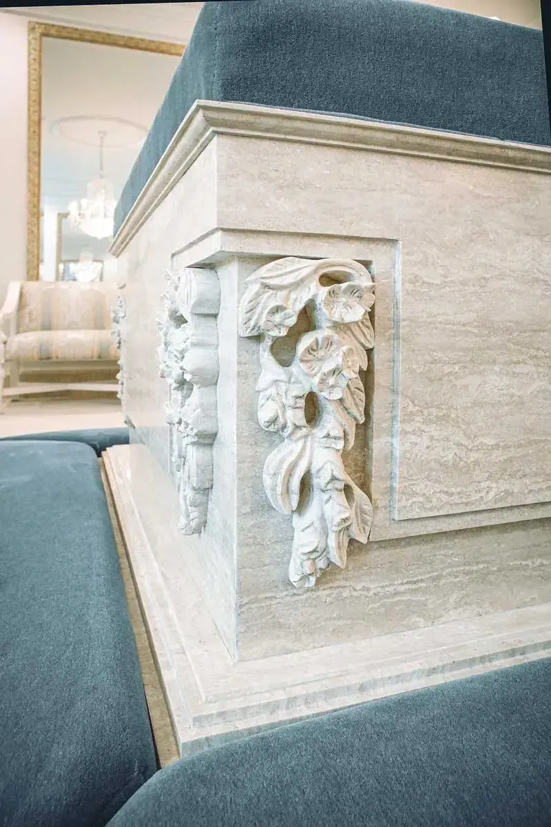 Close view of a carved flower relief on the corner of a travertine base beside blue upholstered cushions.