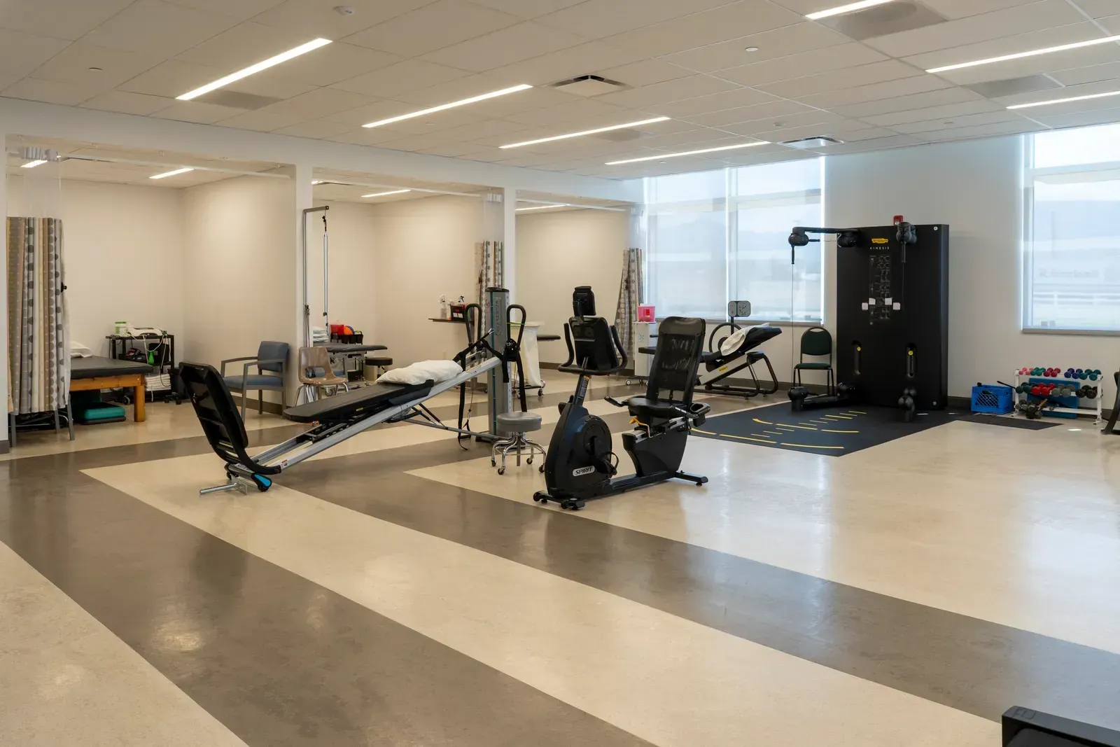 Therapy gym with exercise equipment and curtained treatment areas on a striped floor near large windows.