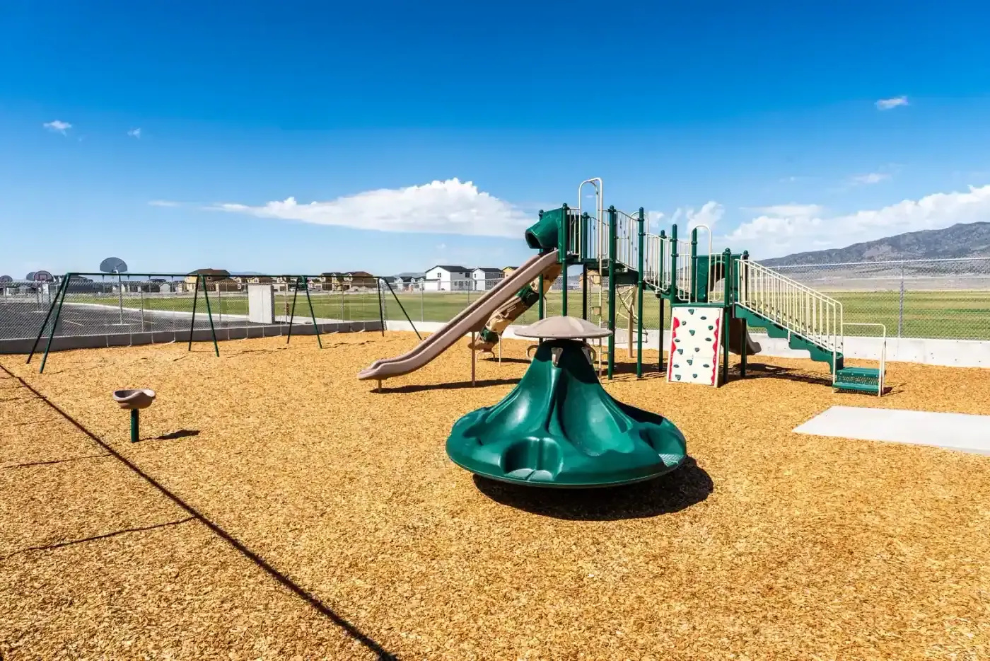 Playground with a green slide structure and a spinner on wood chips, with a fence and mountains in the distance.