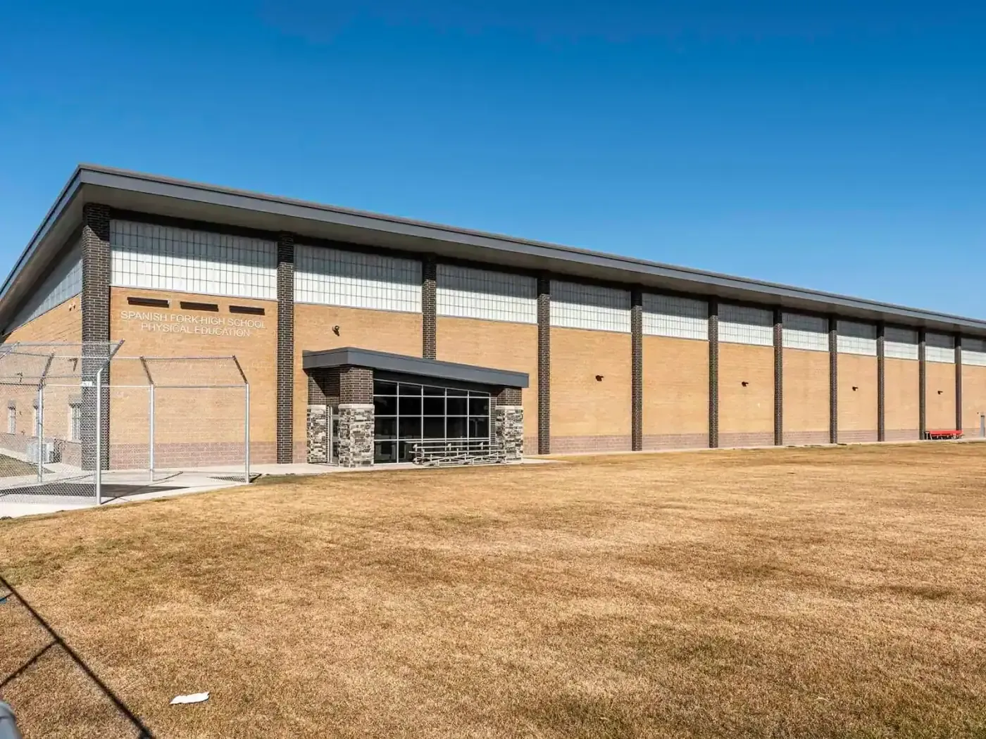 Exterior of a long tan brick building with a glass entrance and a dry grass field in front.