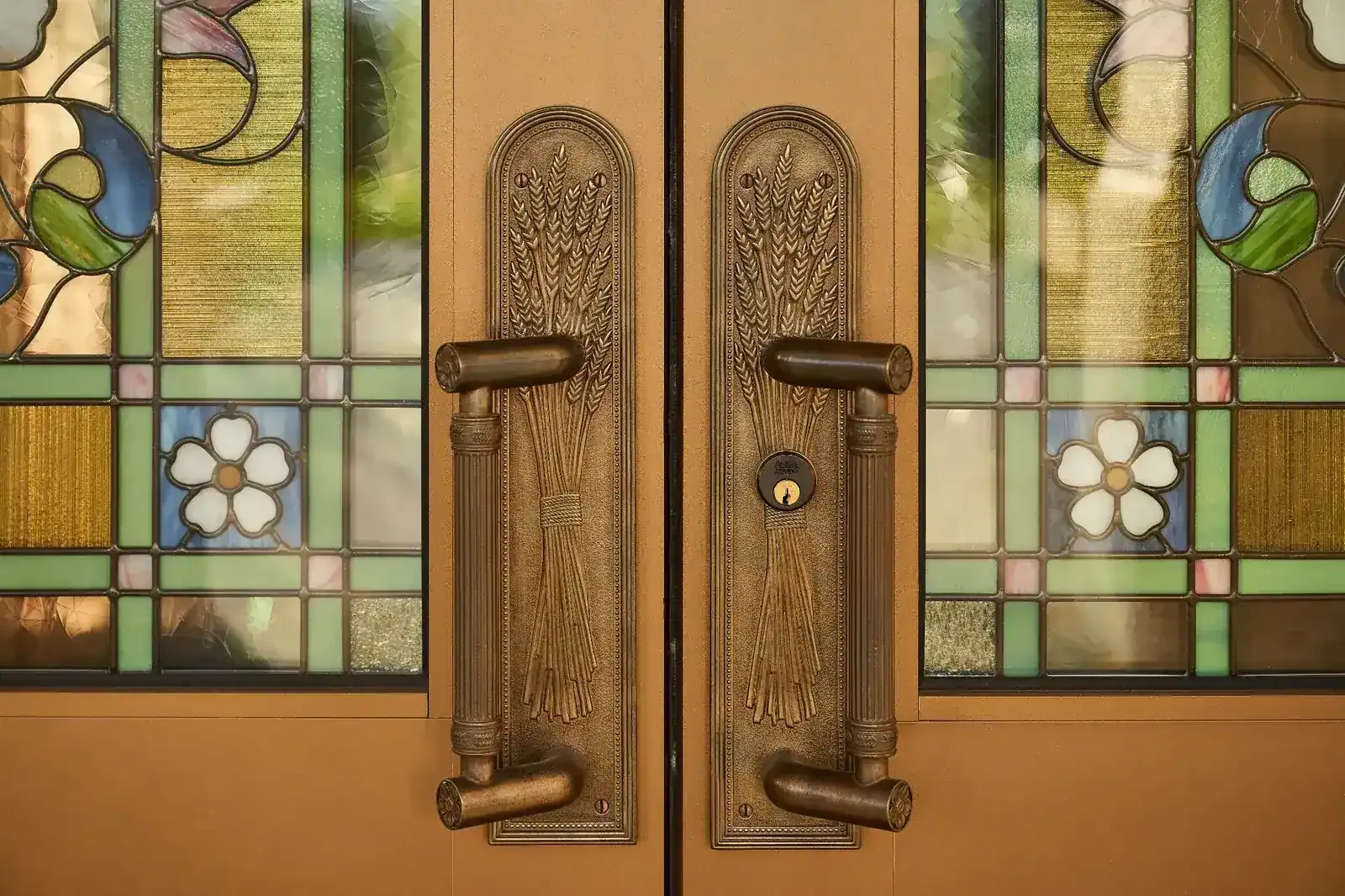 Close view of bronze door handles with a sheaf-of-wheat design beside stained-glass panels with white flowers