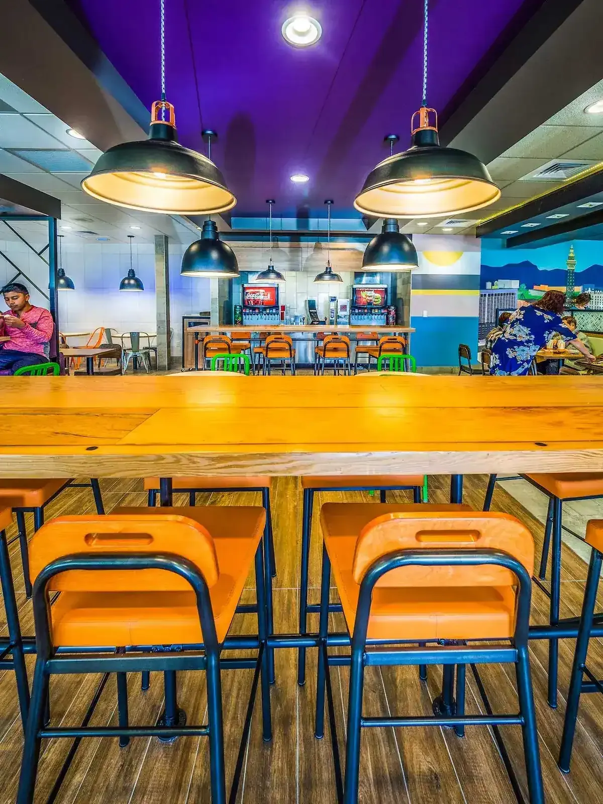 Row of orange metal stools at a long wood table with pendant lamps overhead.